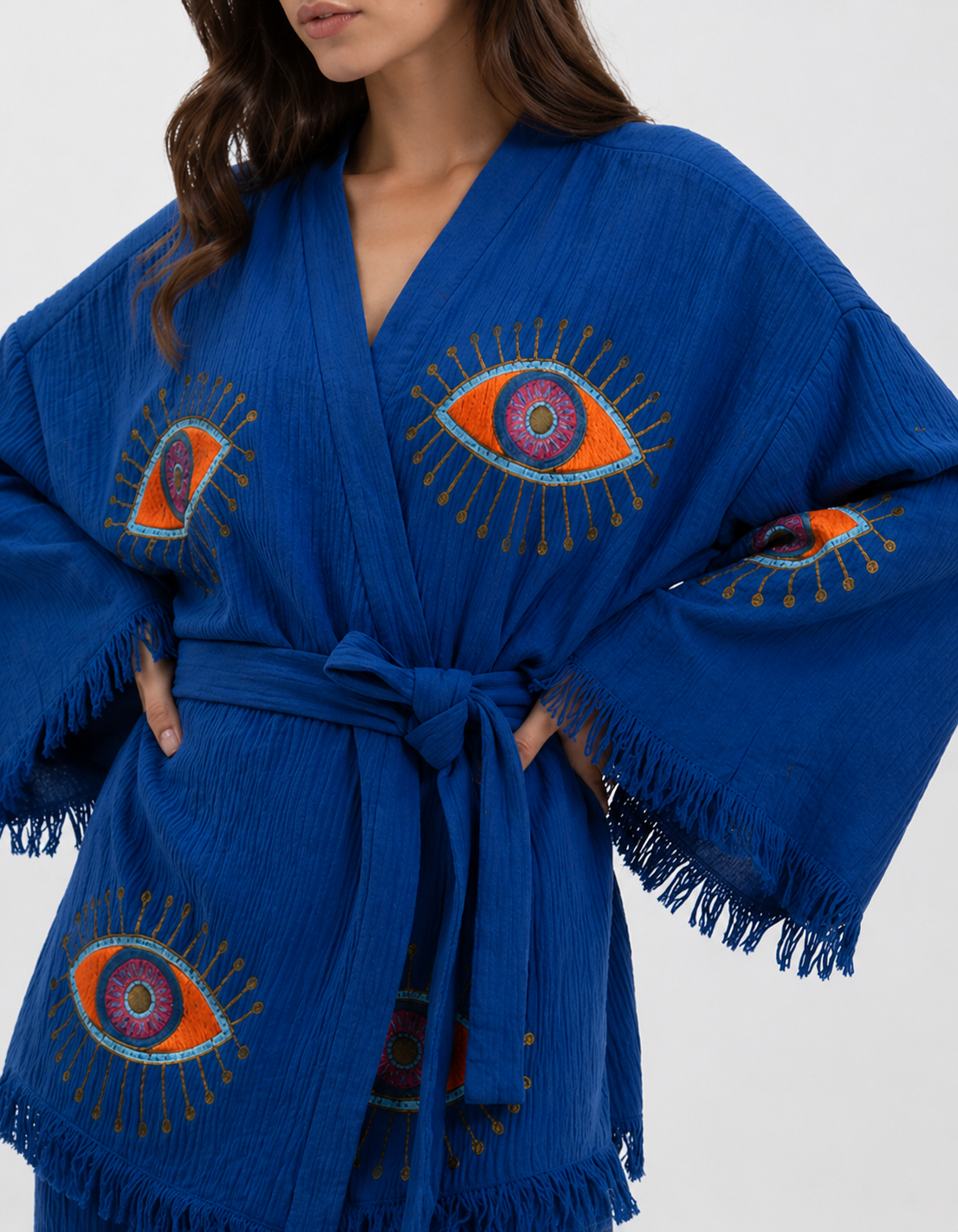 Close-up of a cobalt blue textured cotton kimono showing embroidered-style evil eye motifs in orange, pink, turquoise, and gold, a wrap neckline, matching waist tie, wide sleeves, and fringe edging.