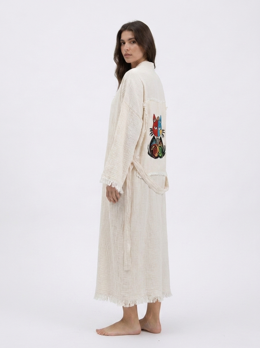 Side view of a cream cotton kimono robe showing the colorful embroidered cat design on the back, lightweight textured fabric, matching waist belt, wide sleeves, and delicate fringe trim.