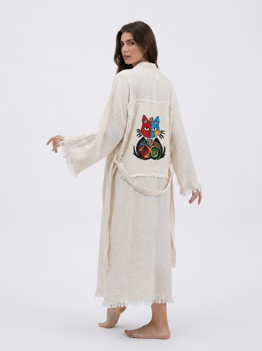 Back-side view of a cream textured cotton kimono robe featuring a colorful embroidered cat patch, wide fringed sleeves, a matching waist belt, and a relaxed ankle-length silhouette.