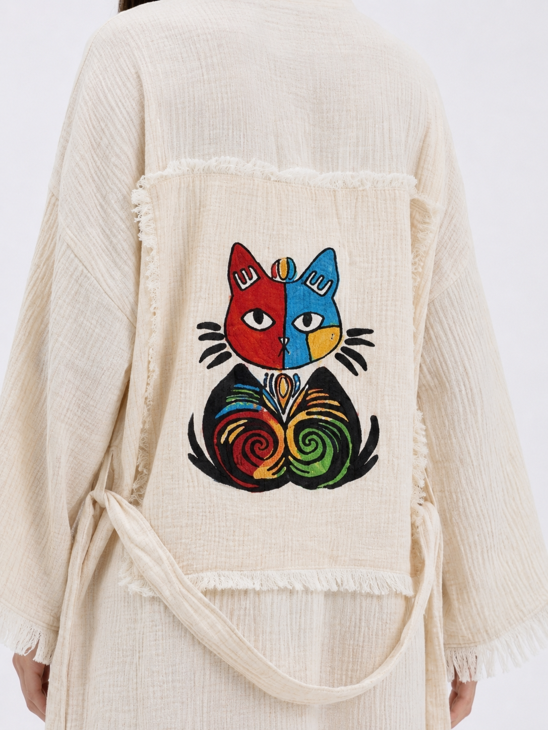 Close-up of the colorful embroidered cat motif on the back of a cream cotton kimono robe, showing red, blue, yellow, green, and black stitching on a textured patchwork panel with raw fringe edges.