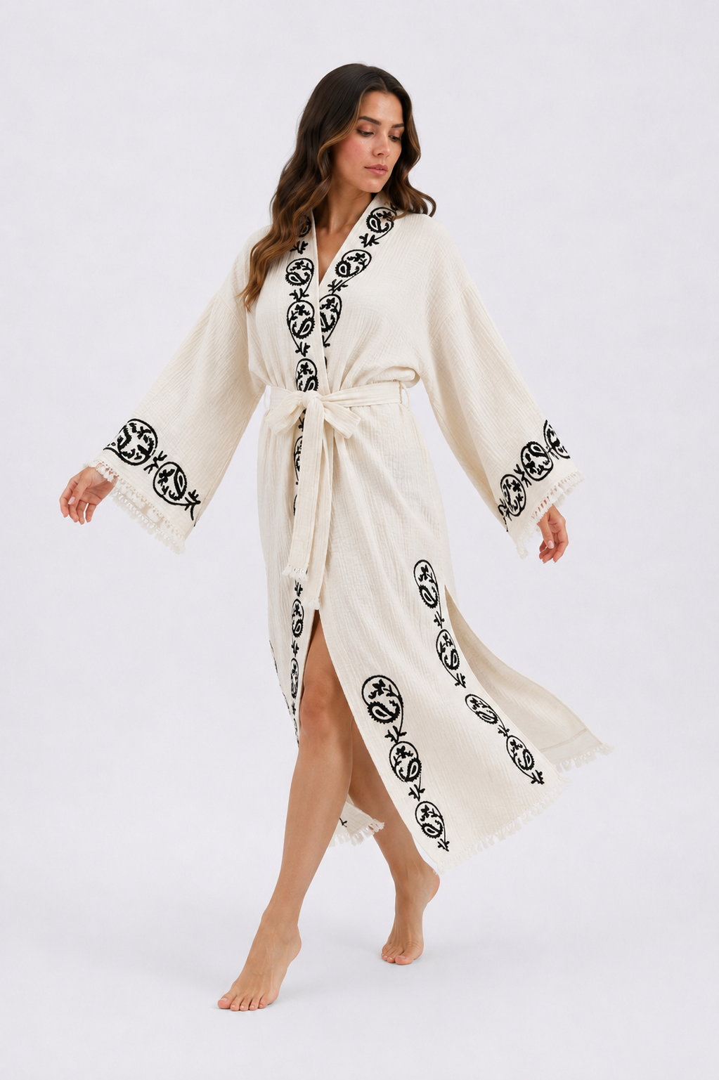 Woman walking in a flowing cream cotton kimono robe featuring handmade black wood block printed botanical medallion motifs along the collar, sleeves, and front panels, with wide sleeves, a tied waist, side openings, and fringed hem.