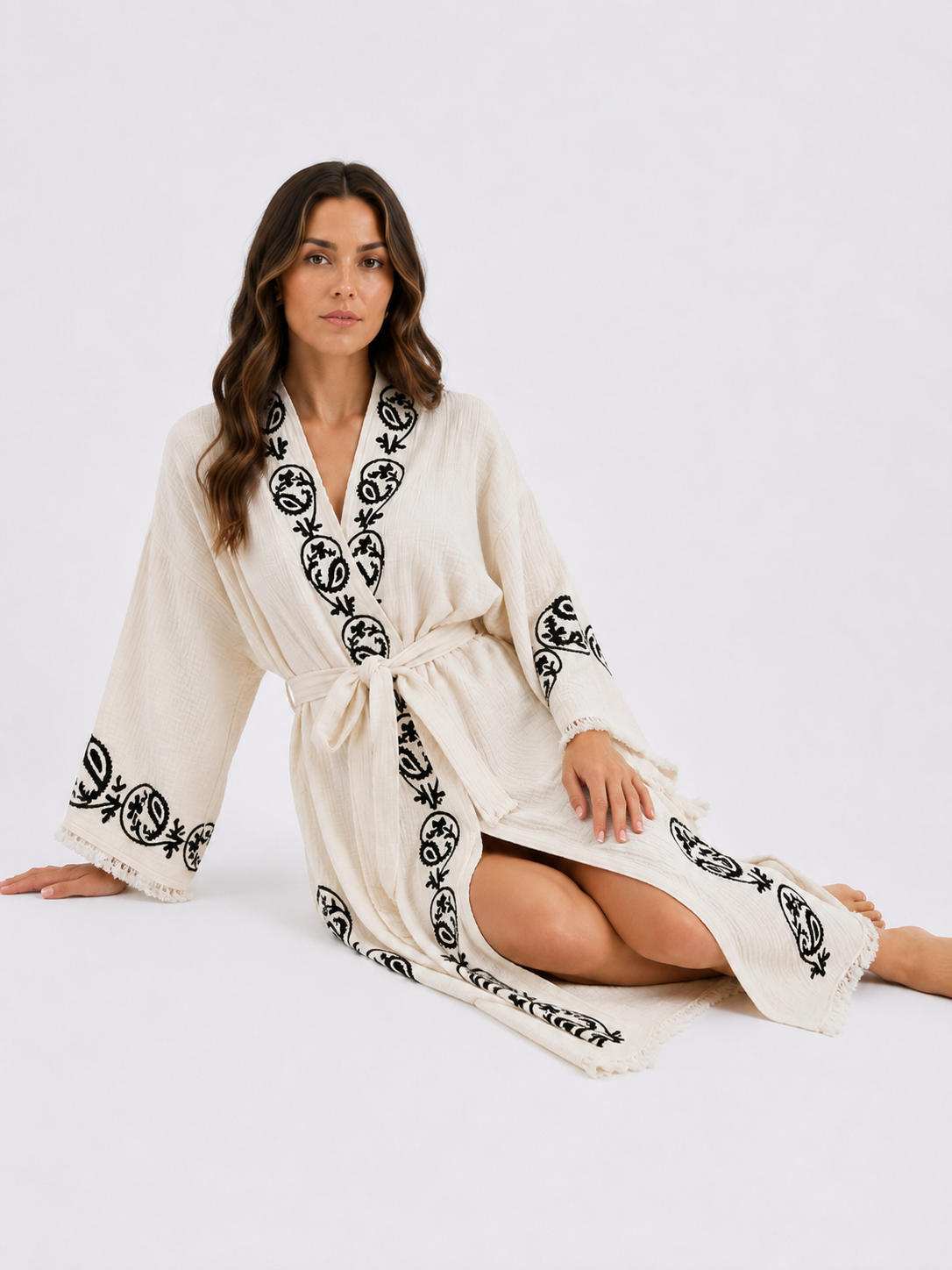 Woman seated on the studio floor wearing a cream textured cotton kimono robe with black handmade wood block printed botanical motifs, wide sleeves, matching waist belt, front opening, side slit, and soft fringe detailing.