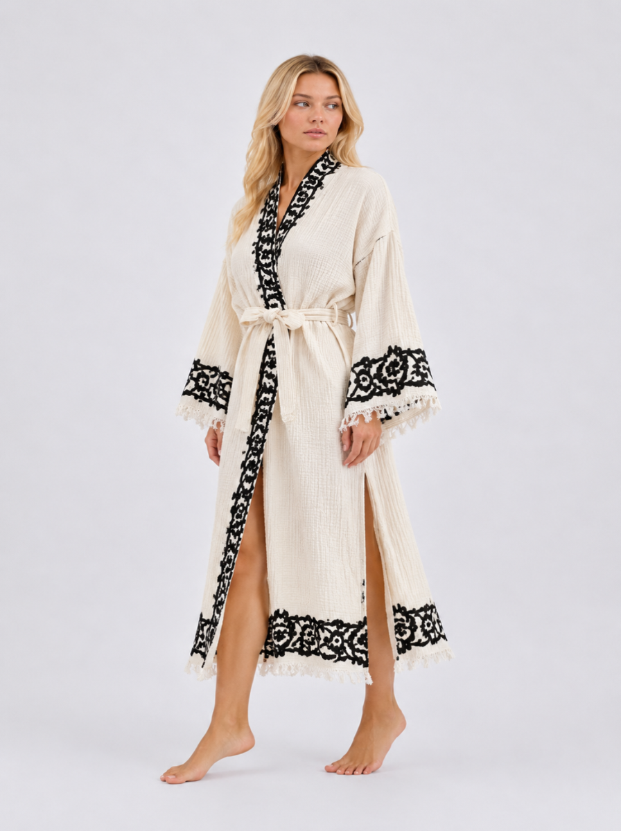 Front view of a woman wearing a long cream textured kimono robe with contrasting black ornamental hand printed along the shawl collar, wide sleeves, front opening, and fringed ankle-length hem, finished with a matching waist belt.