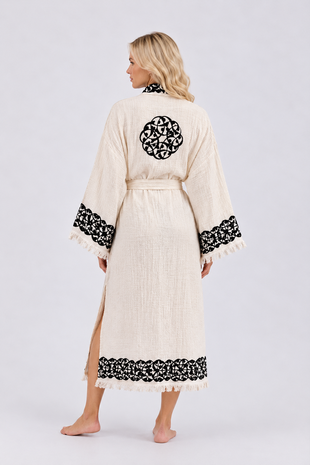 Back view of a cream textured kimono robe featuring a circular black hand printed medallion across the upper back, ornamental wood block printed along the wide sleeve cuffs and hem, a matching waist tie, side slits, and delicate fringe trim.
