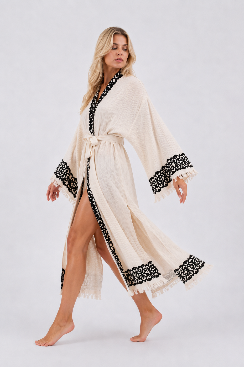 Woman walking in a flowing cream textured kimono robe with bold black hand printed borders, wide bell sleeves, a tied waist, deep side openings, and fringe-trimmed hem, showing the lightweight drape and movement of the fabric.