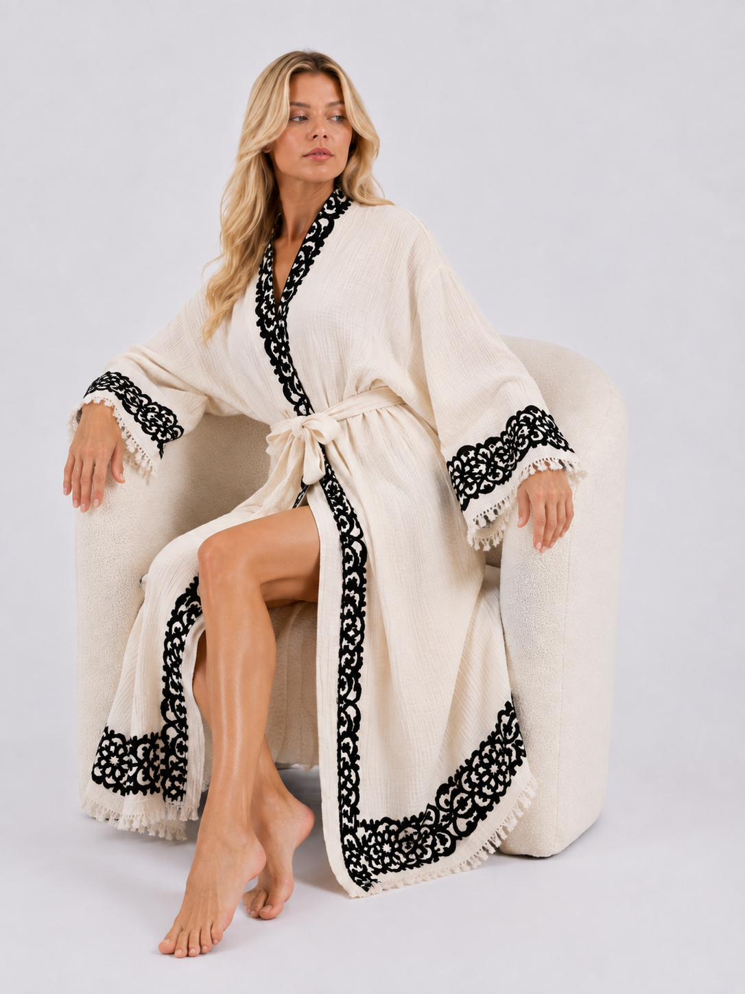 Woman seated in a minimalist studio wearing a cream ankle-length kimono robe with black ornamental print around the collar, cuffs, front opening, and hem, styled with wide sleeves, a matching waist belt, side slit, and soft fringe trim.