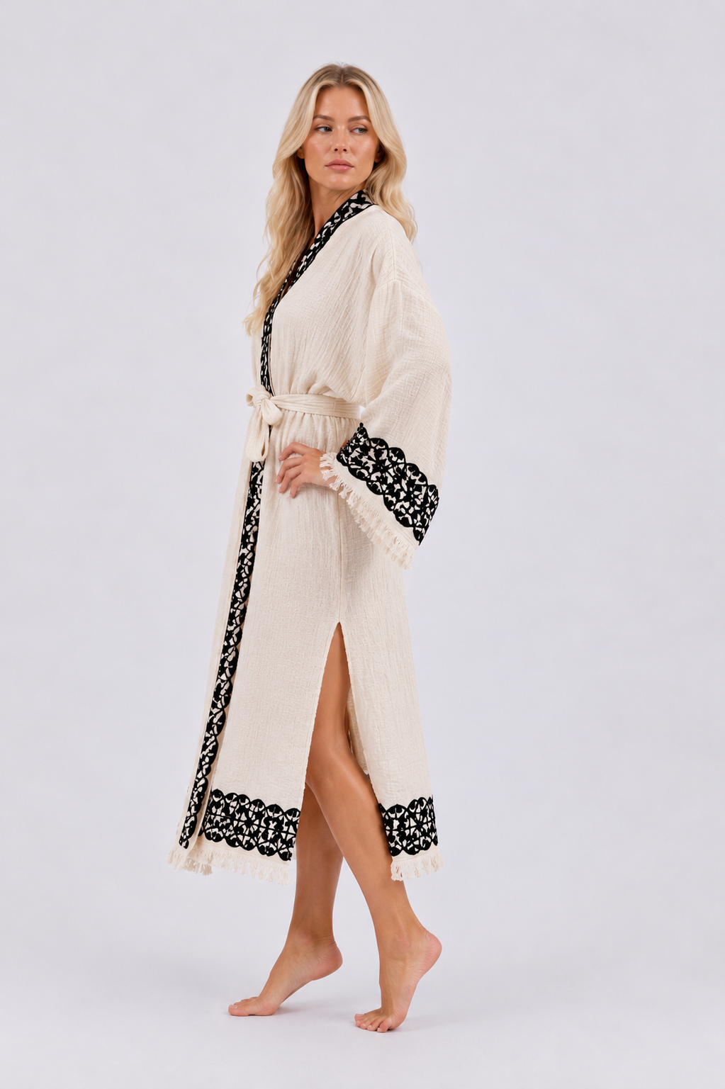 Side view of a long cream kimono robe with black ornamental wood block printed framing the collar, cuffs, front opening, and lower hem, showing the relaxed wrap silhouette, wide sleeves, matching fabric belt, side slit, and fringe detailing.