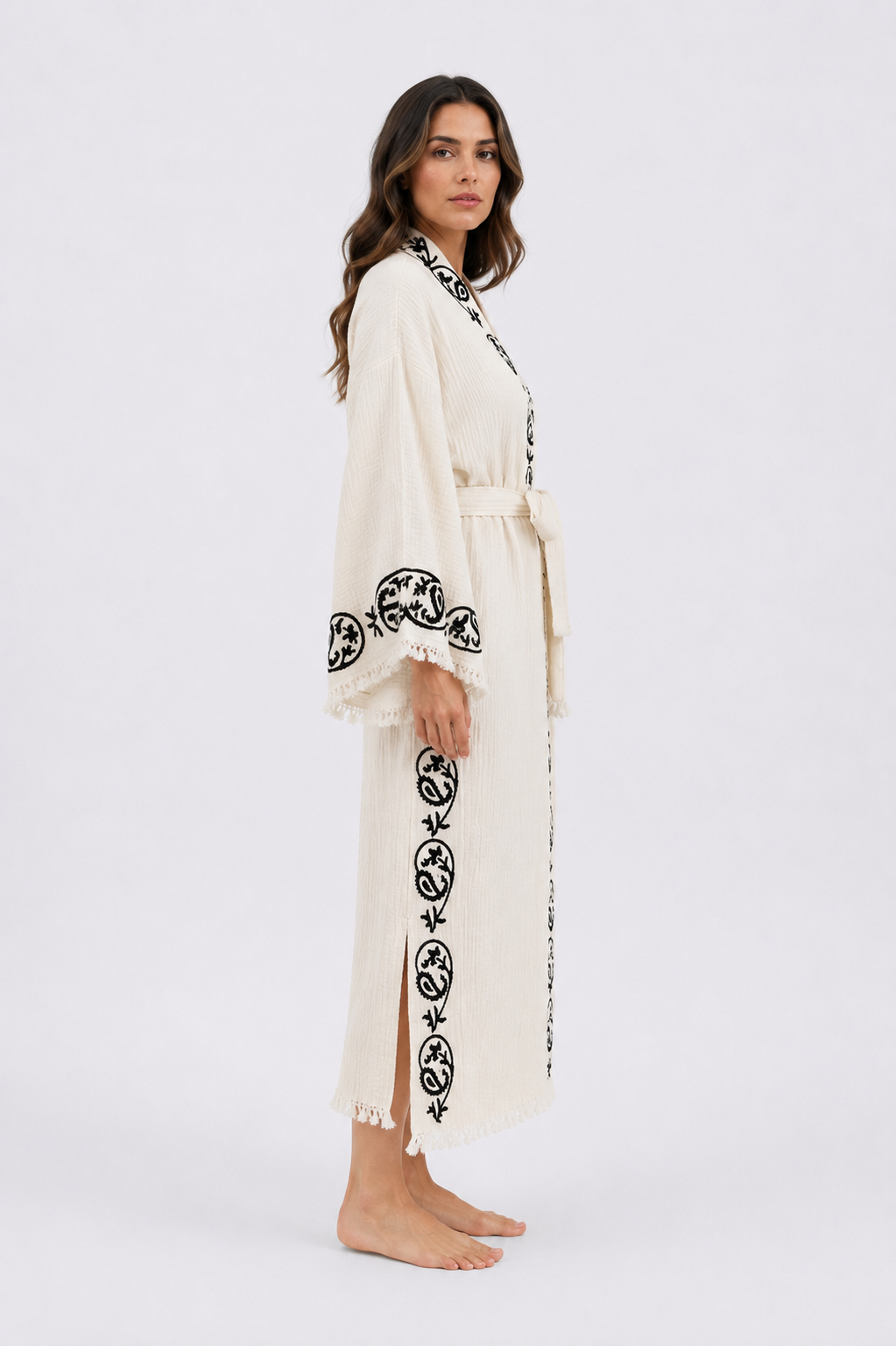 Side view of a woman wearing a cream textured kimono robe with handmade black wood block printed motifs along the collar, wide sleeve cuffs, front border, and side panel, styled with a matching waist tie, ankle-length silhouette, and fringe edging.