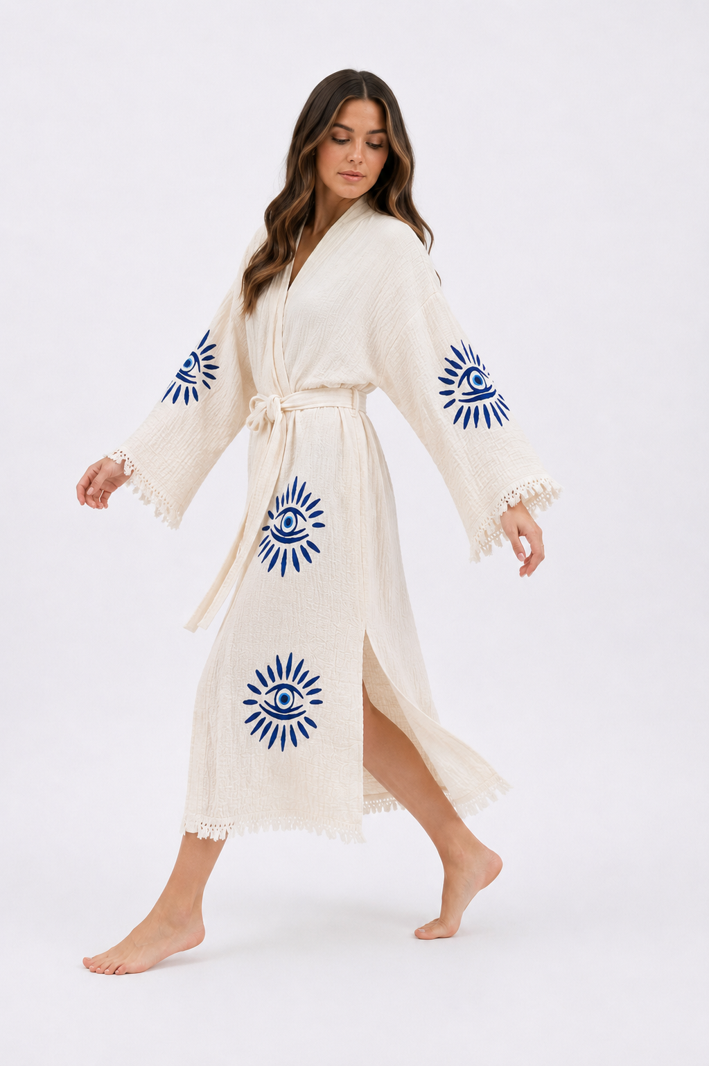 Woman walking in a cream cotton kimono robe decorated with cobalt blue evil eye motifs, showing the flowing wrap design, side slit, wide sleeves, tied waist, textured fabric, and fringe-trimmed hem in motion.