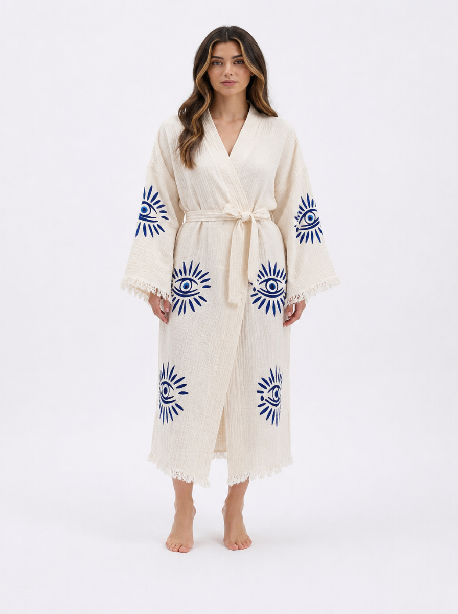 Front view of a woman wearing a cream textured cotton kimono robe with cobalt blue evil eye embroidery, wide sleeves, a matching waist belt, ankle-length silhouette, and delicate fringe trim along the cuffs and hem.