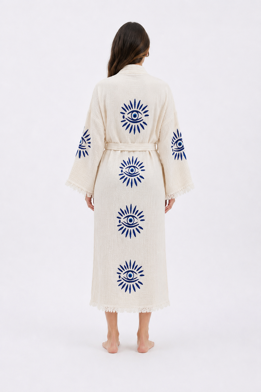 Back view of a cream cotton kimono robe featuring cobalt blue evil eye motifs arranged vertically across the back and sleeves, with a tied waist, relaxed fit, wide sleeves, and soft fringed edges.