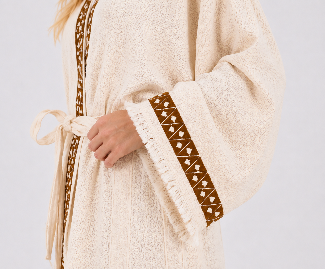 Close-up of a cream textured cotton kimono robe showing brown geometric cuff and neckline trim, a matching waist tie, wide sleeve, natural woven texture, and cream fringe edging.