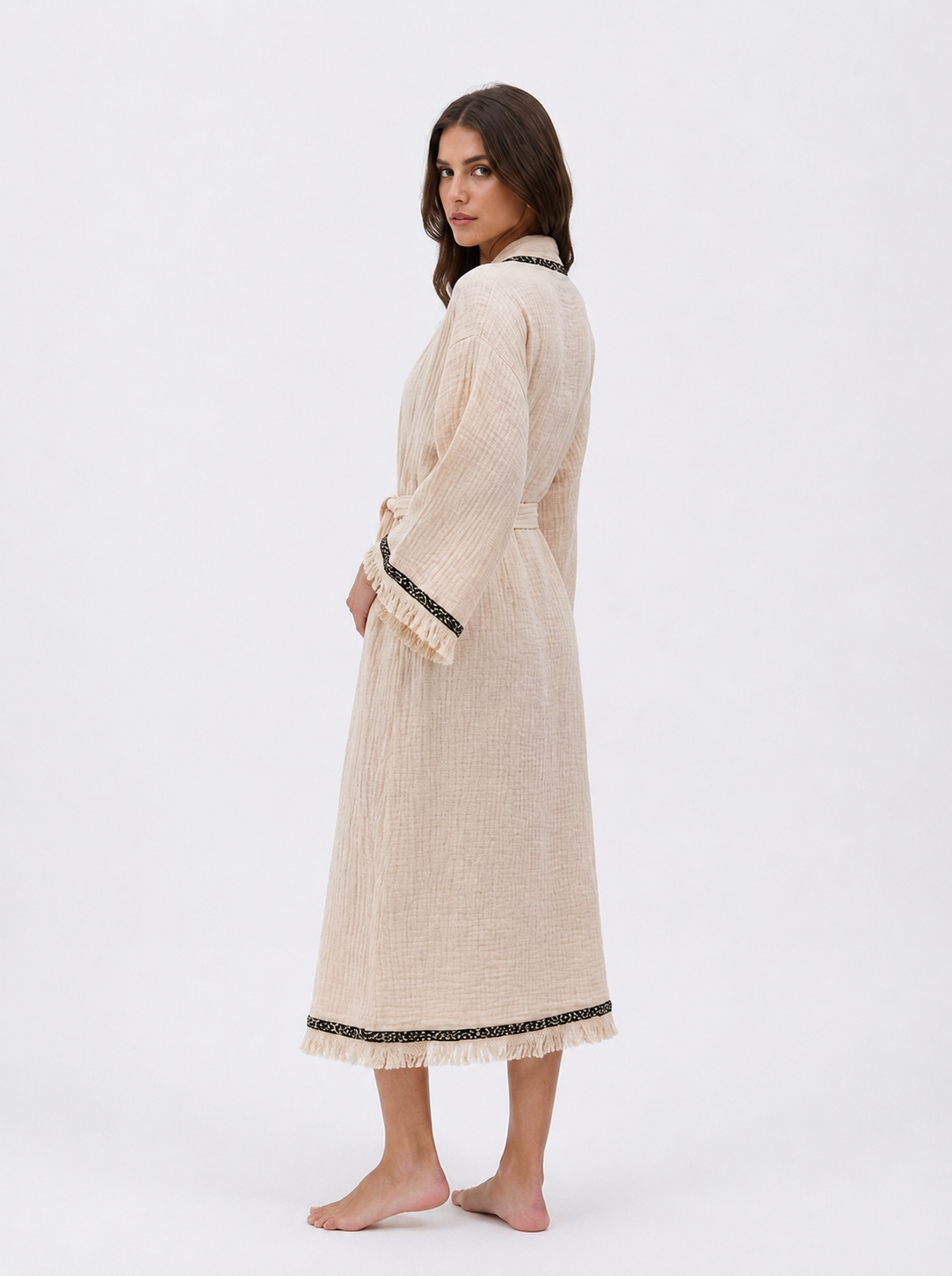 Side-back view of a long cream cotton kimono robe with a belted waist, wide sleeves, black patterned trim, softly textured natural fabric, and fringe edging along the cuffs and hem.