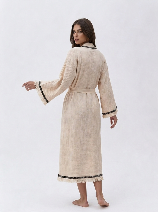 Back view of a cream textured cotton kimono robe showing the relaxed long silhouette, matching tie belt, wide sleeves, black patterned border trim, and soft fringe along the cuffs and hem.