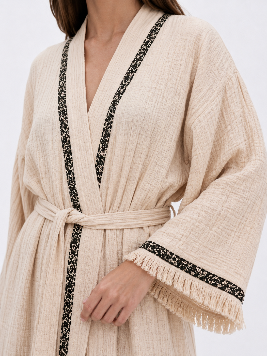 Close-up of a cream cotton kimono robe showing the soft crinkled fabric texture, black patterned neckline trim, matching waist tie, wide sleeve, and delicate fringe cuff detail.
