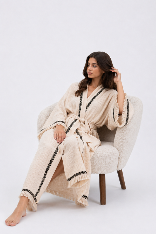 Seated model wearing a cream textured cotton kimono robe with black patterned trim, a matching waist belt, wide fringed sleeves, and a relaxed full-length silhouette.