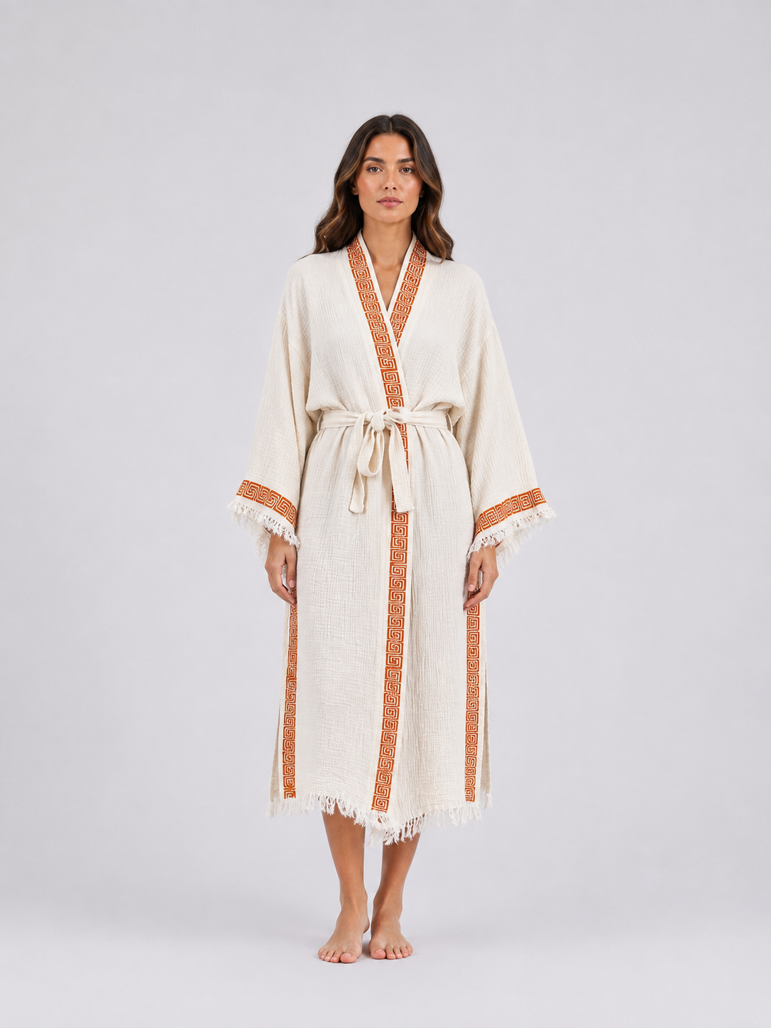 Front view of a model wearing a long cream textured cotton kimono robe with burnt-orange geometric trim, wide sleeves, cream fringe edging, a matching waist belt, and a relaxed ankle-length fit.