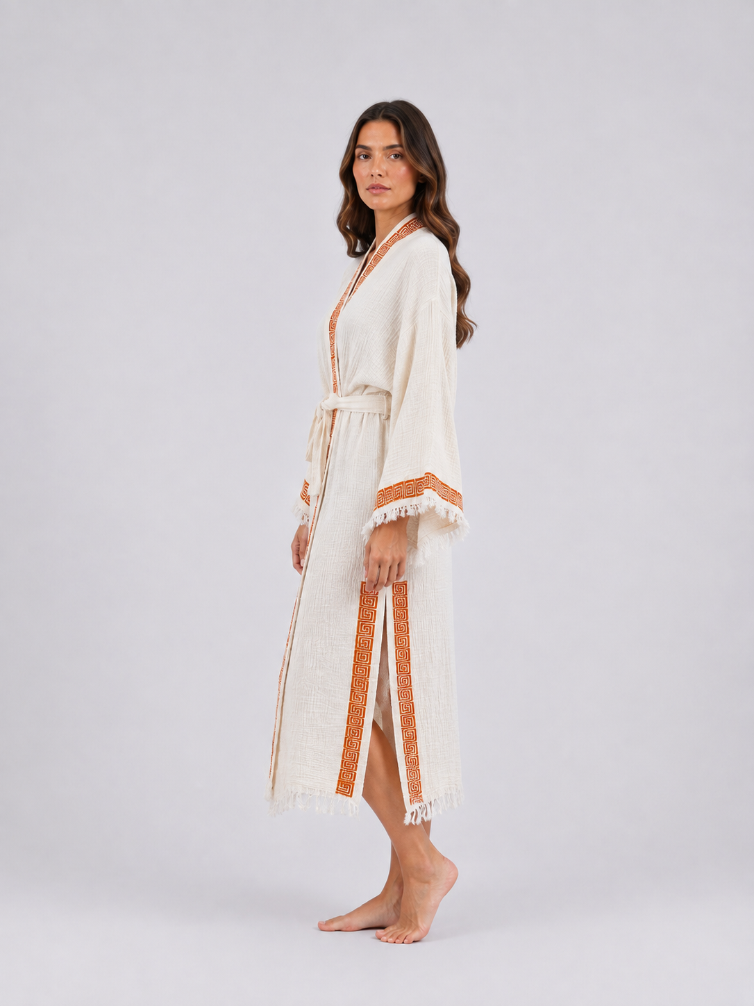 Side view of a cream cotton kimono robe showing the flowing ankle-length silhouette, burnt-orange geometric border, side opening, matching tie belt, wide sleeves, and fringed cuffs and hem.