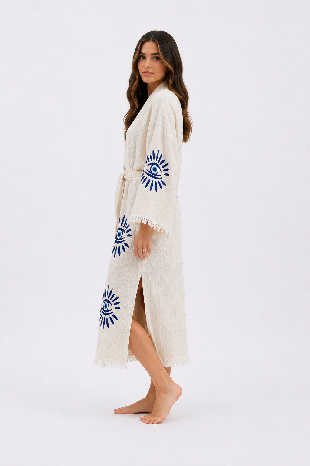 Side view of a cream textured kimono robe with blue evil eye embroidery on the sleeve and lower body, showing the wrap silhouette, matching fabric belt, side opening, ankle length, and fringed cuffs.