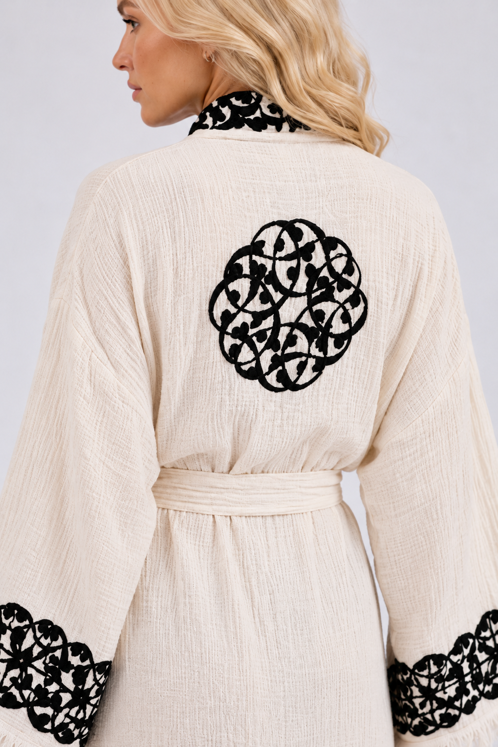 Close-up of the back of a cream textured kimono robe showing an intricate circular black hand printed medallion, coordinating black floral embroidery on the collar and wide sleeve cuffs, a matching waist belt, and visible woven fabric texture.