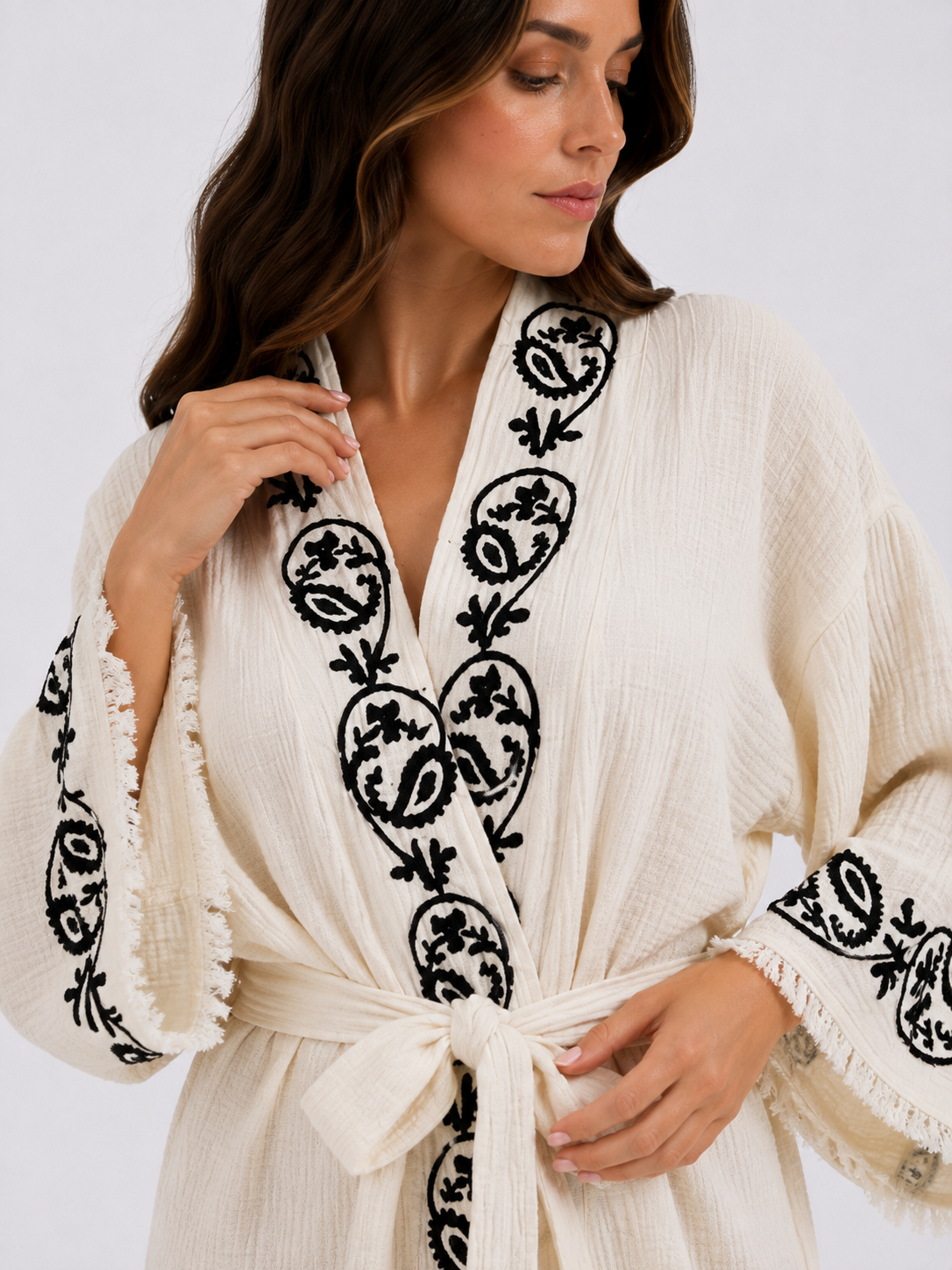 Close-up of a cream textured kimono robe showing handmade black wood block printed botanical motifs along the shawl collar and wide sleeve cuffs, with a matching fabric belt, natural woven texture, and hand-finished fringe trim.