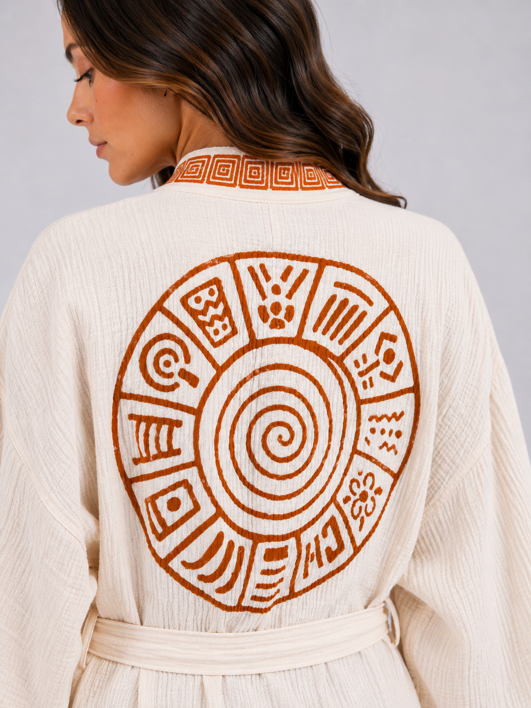 Close-up back view of a cream cotton kimono robe featuring a large burnt-orange circular spiral design with surrounding geometric symbols, a patterned collar, textured fabric, and matching waist belt.