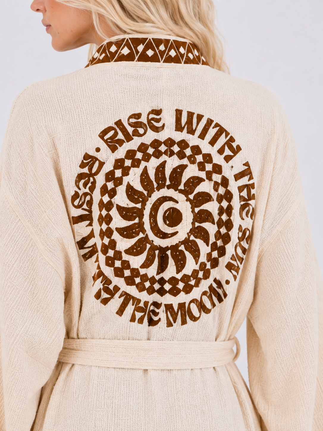 Close-up of the back of a cream cotton kimono robe featuring a brown sun and crescent moon design with the words Rise with the Sun, Rest with the Moon, plus geometric collar trim.