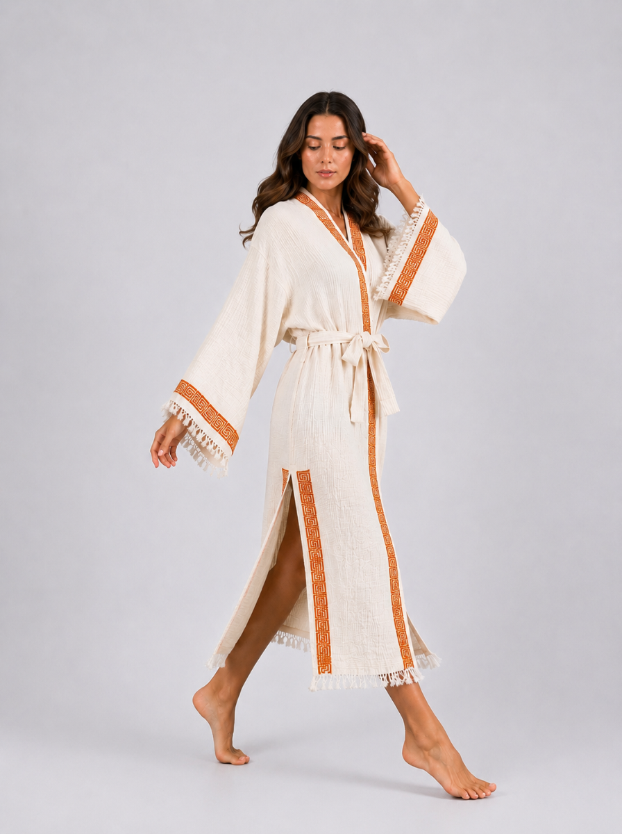 Model walking in a long cream cotton kimono robe with burnt-orange geometric trim, showing the flowing fabric, side opening, wide fringed sleeves, matching belt, and relaxed movement.