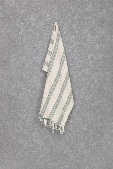 Gelin Turkish cotton peshtemal towel hanging against a gray wall, showing its ivory weave, striped pattern and tassel fringe.