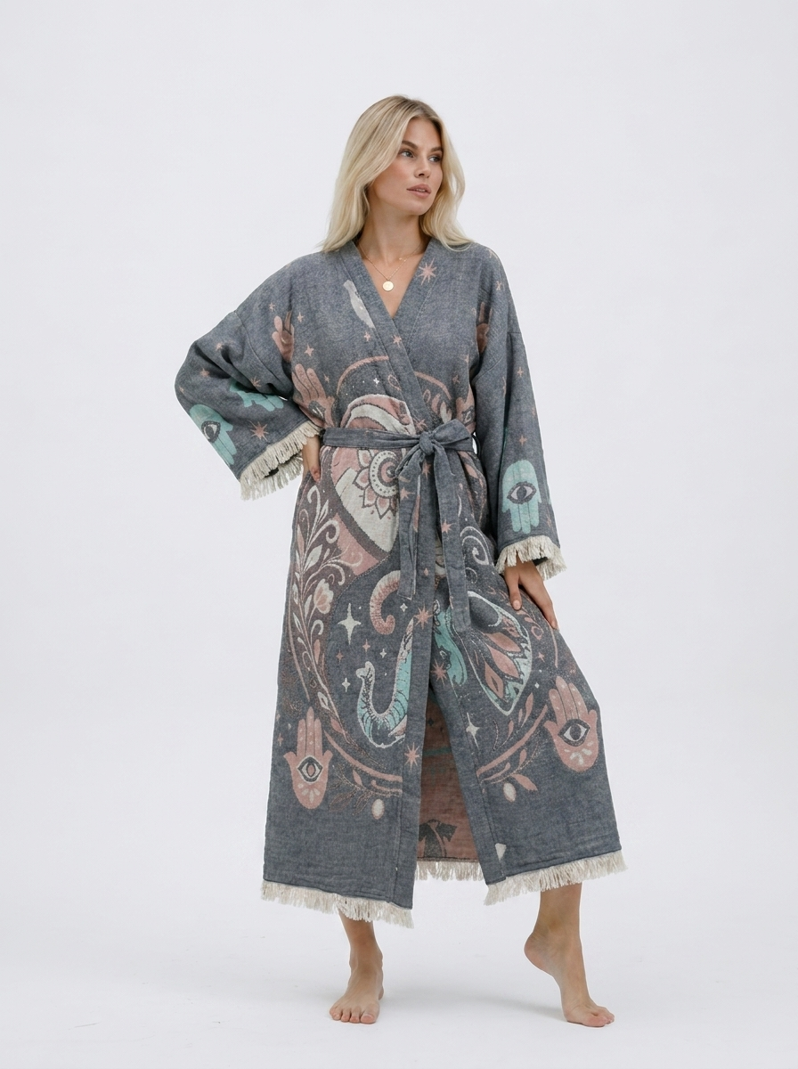 Front view of a blonde model wearing a long grey jacquard cotton kimono robe with pastel elephant, Hamsa hand, evil eye, and celestial motifs, a matching waist belt, wide sleeves, and cream fringe trim.