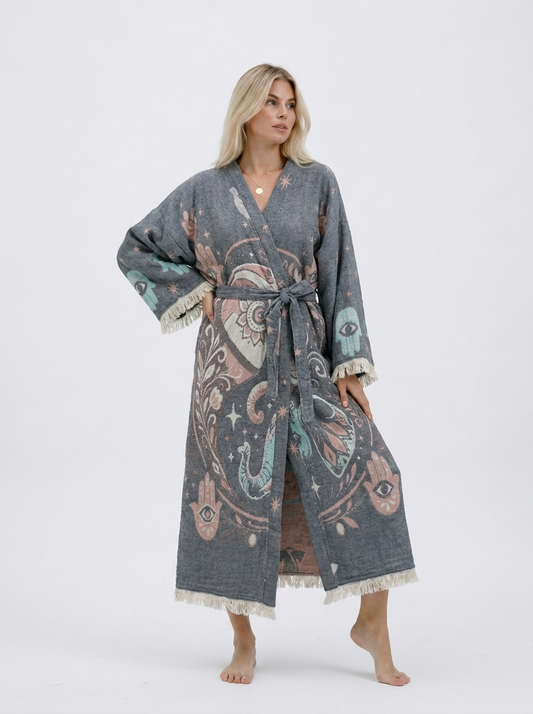 Front view of a blonde model wearing a long grey jacquard cotton kimono robe with pastel elephant, Hamsa hand, evil eye, and celestial motifs, a matching waist belt, wide sleeves, and cream fringe trim.