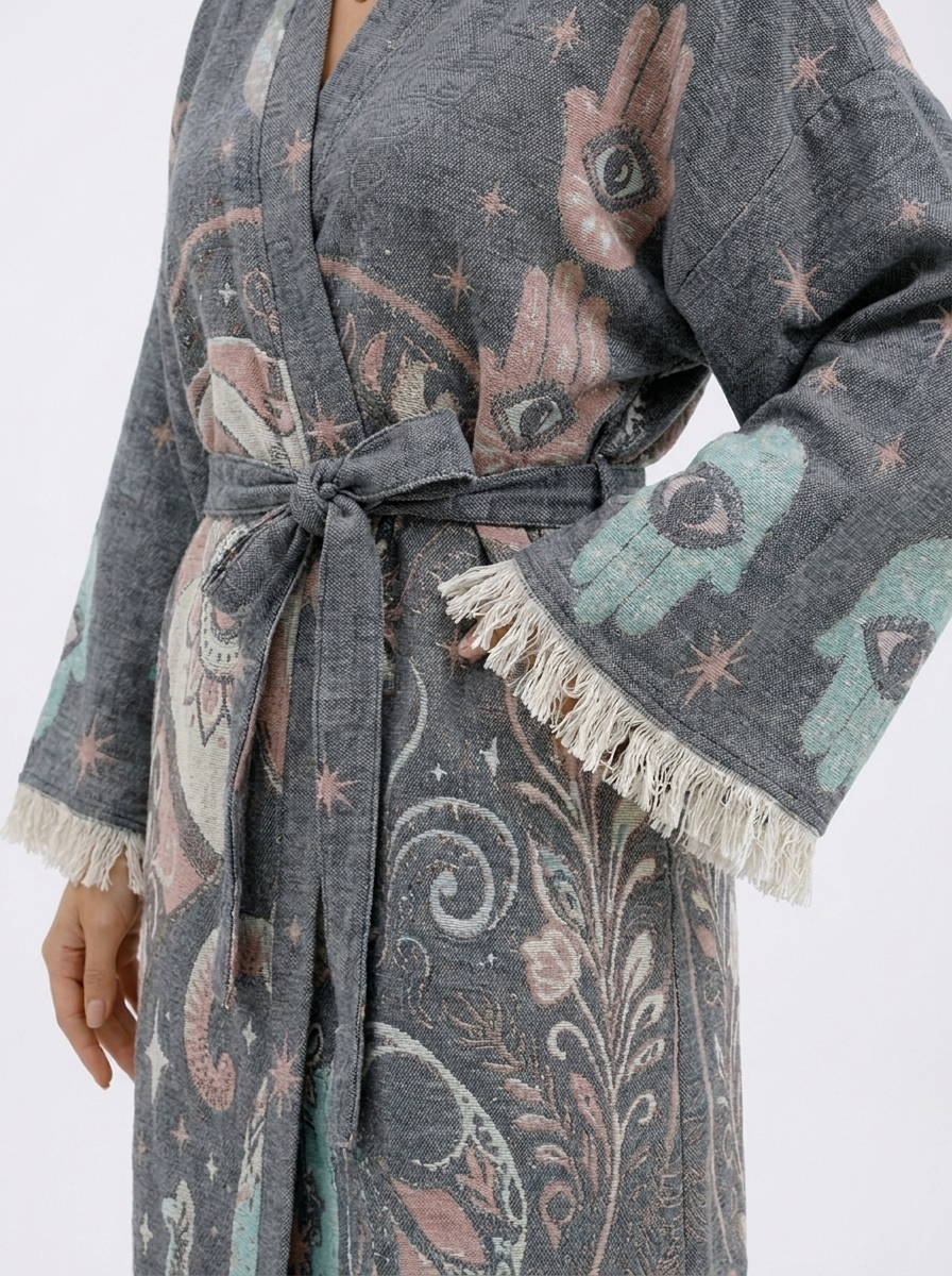 Close-up of a grey jacquard cotton kimono robe showing the textured woven fabric, pastel Hamsa hand and evil eye motifs, matching waist tie, wide sleeve, and cream fringe edging.