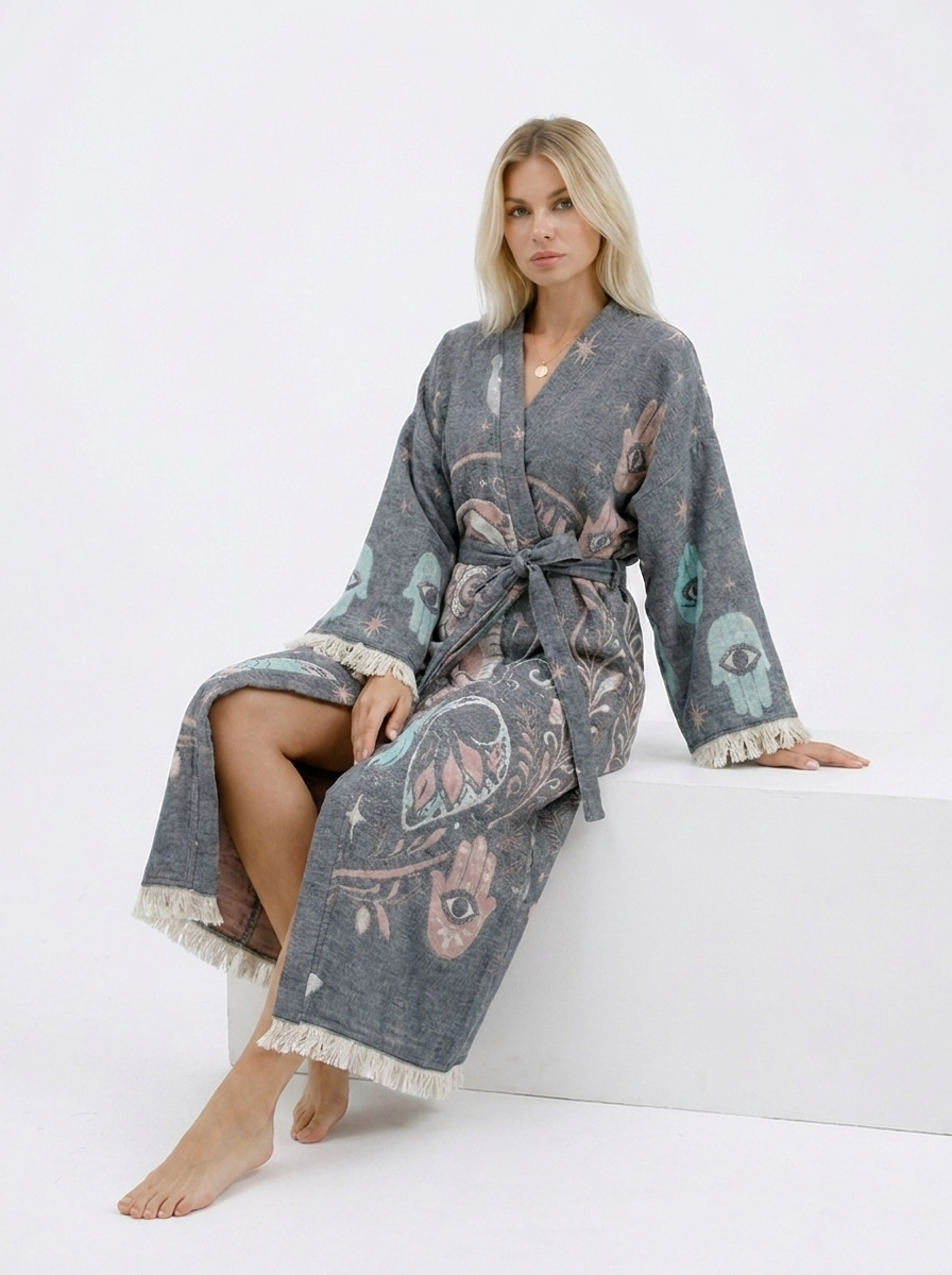 Seated model wearing a grey jacquard cotton kimono robe with pastel elephant, Hamsa hand, evil eye, and celestial designs, a relaxed ankle-length fit, wide sleeves, belt, and fringe trim.