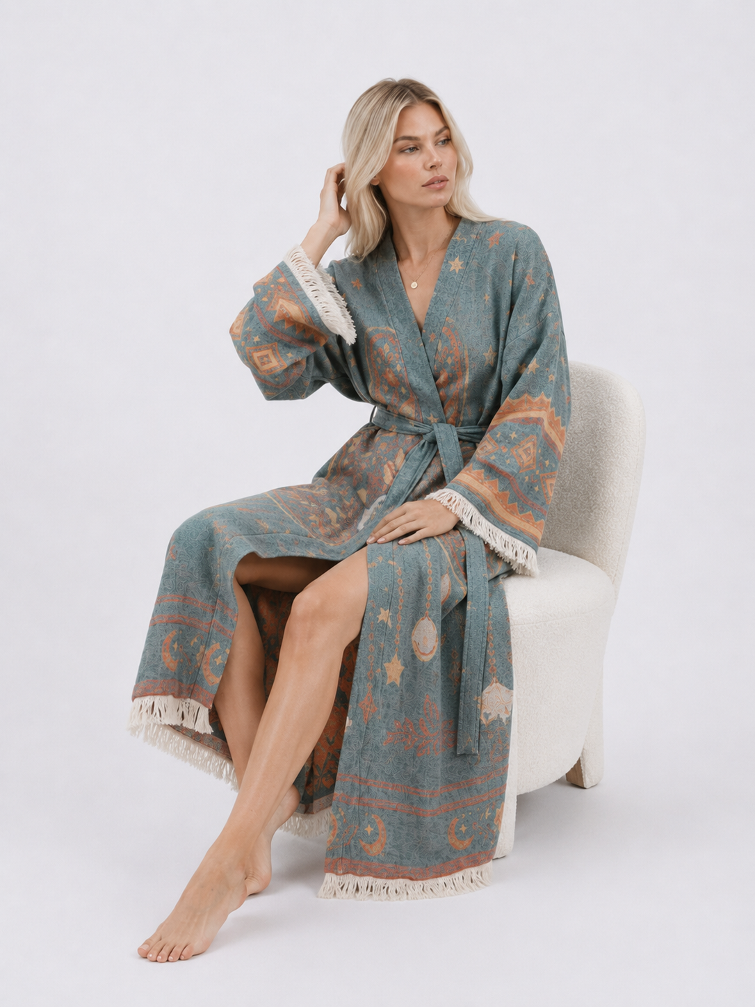 Seated model wearing a teal jacquard cotton kimono robe with Hamsa hand, evil eye, star, and celestial motifs, a belted waist, wide fringed sleeves, and a relaxed ankle-length fit.