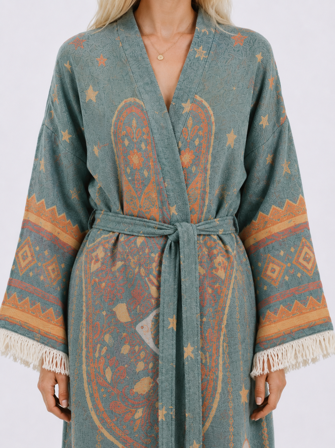 Close-up of a teal jacquard cotton kimono robe showing the woven Hamsa-inspired pattern, coral and gold celestial motifs, matching tie belt, wide sleeves, and cream fringe trim.