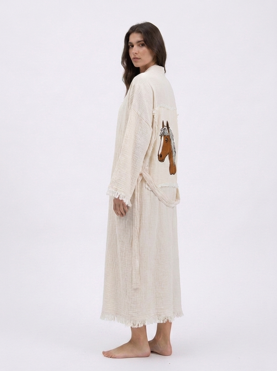 Side-back view of the Happy Horse cream cotton kimono robe featuring a hand-embroidered horse patch, lightweight textured fabric, wide sleeves, matching waist belt, and softly fringed edges.
