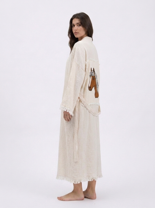 Side-back view of the Happy Horse cream cotton kimono robe featuring a hand-embroidered horse patch, lightweight textured fabric, wide sleeves, matching waist belt, and softly fringed edges.