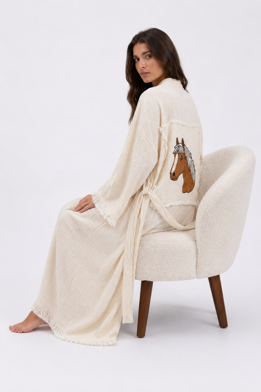 Seated model wearing the Happy Horse cream cotton kimono robe, showing the embroidered horse design on the back, relaxed flowing fit, textured natural fabric, wide fringed sleeves, and matching belt.