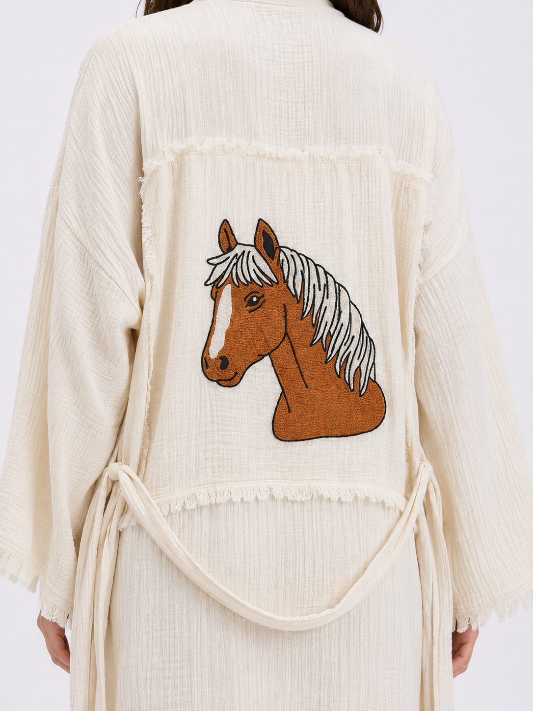 Close-up of the hand-embroidered brown horse motif on the back of a cream natural cotton kimono robe, showing the detailed stitching, textured fabric, patchwork panel, and raw fringe edging.