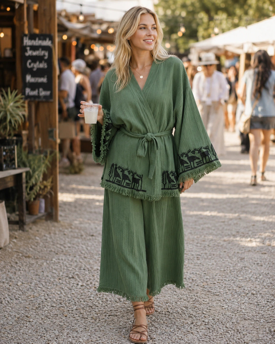 Woman wearing the Nara sage green kimono and matching wide-leg pants set at an outdoor market, with black folk-inspired animal motifs and fringed hems.