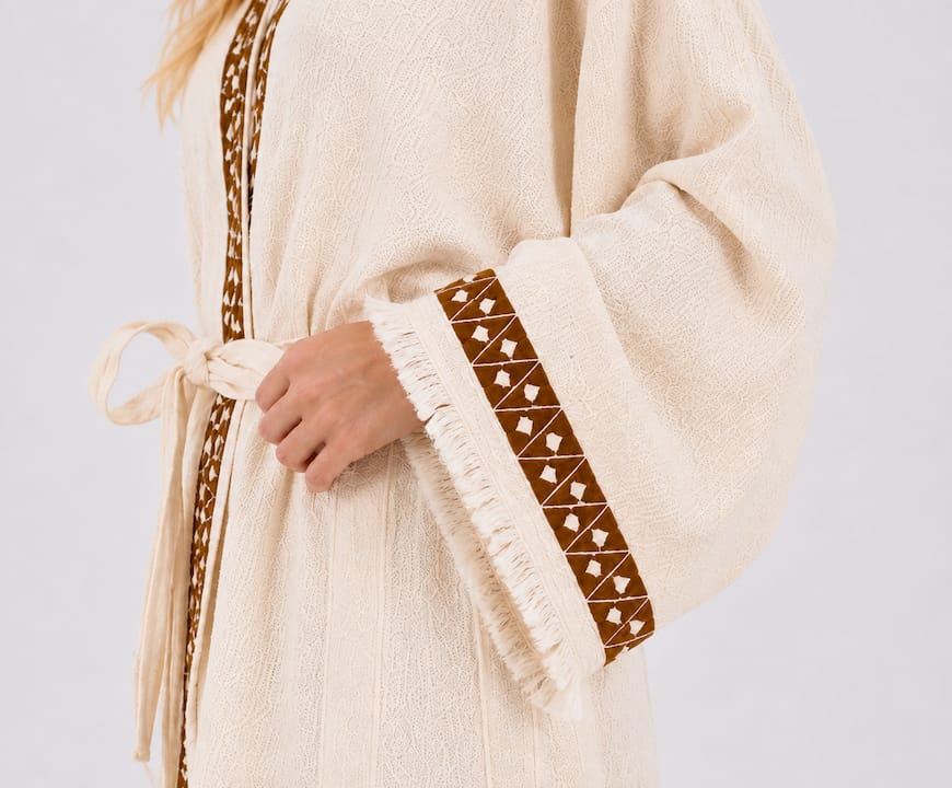 Close-up of a cream cotton kimono robe with brown geometric trim, matching waist tie, wide sleeve, woven texture, and cream fringe.
