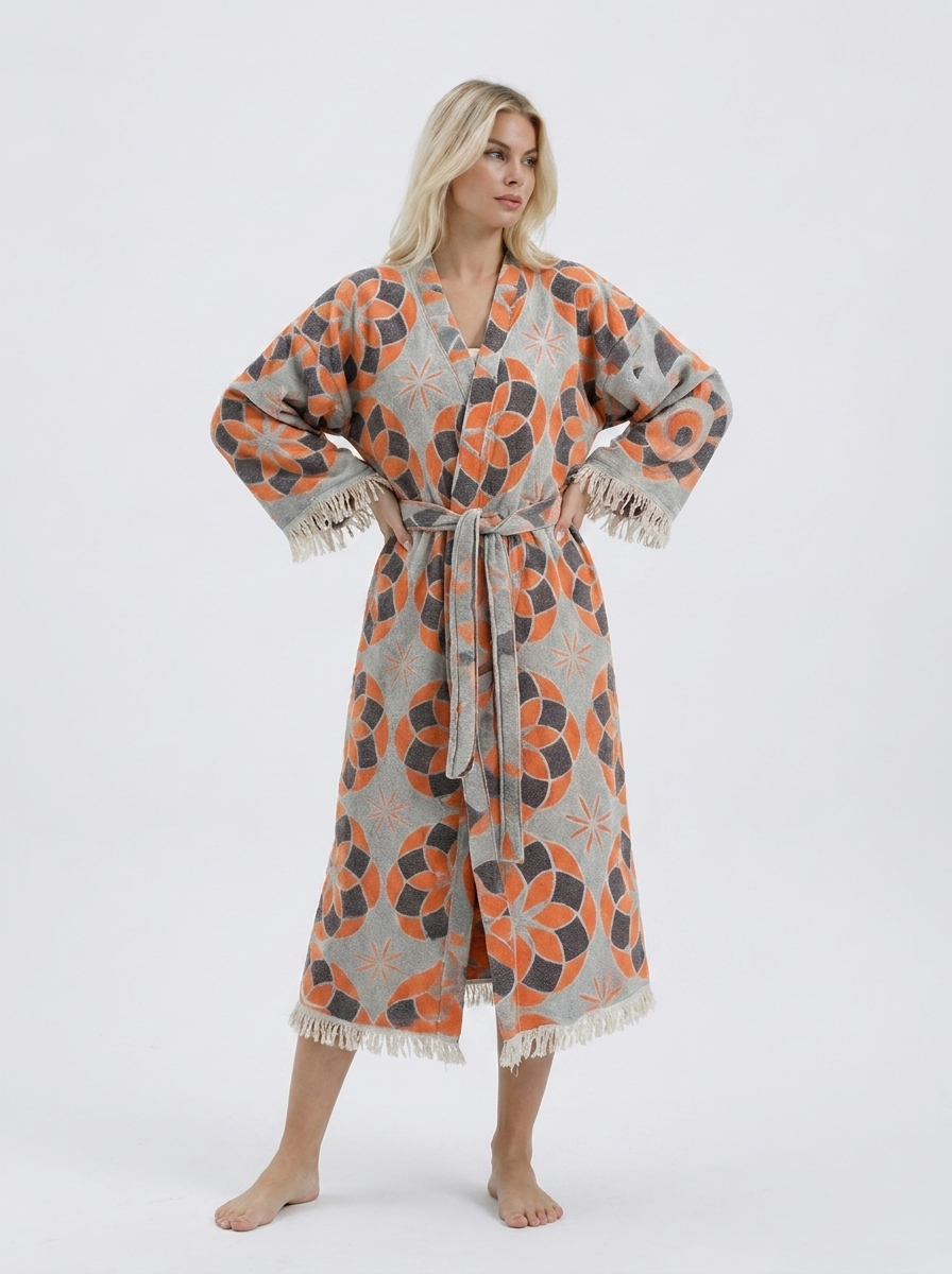 Blonde model wearing a long orange and grey jacquard cotton kimono robe with geometric floral motifs, matching waist belt, wide sleeves, and cream fringe trim.