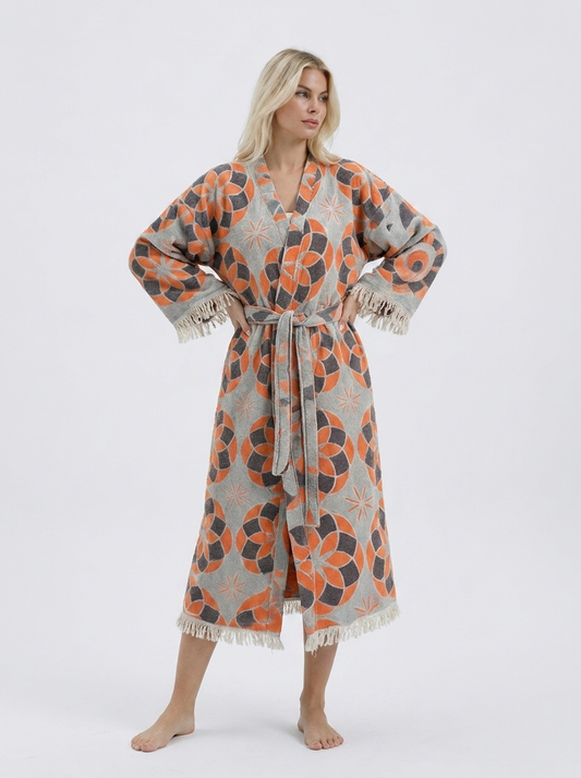 Blonde model wearing a long orange and grey jacquard cotton kimono robe with geometric floral motifs, matching waist belt, wide sleeves, and cream fringe trim.