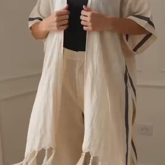 Asya Turkish cotton kimono shown in motion on a woman, featuring lightweight natural ivory woven cotton, an open-front relaxed silhouette, short sleeves with charcoal stripe details, high side splits, flowing drape, and hand-finished tassel fringe. Designed as a beach cover-up and versatile everyday layering piece.