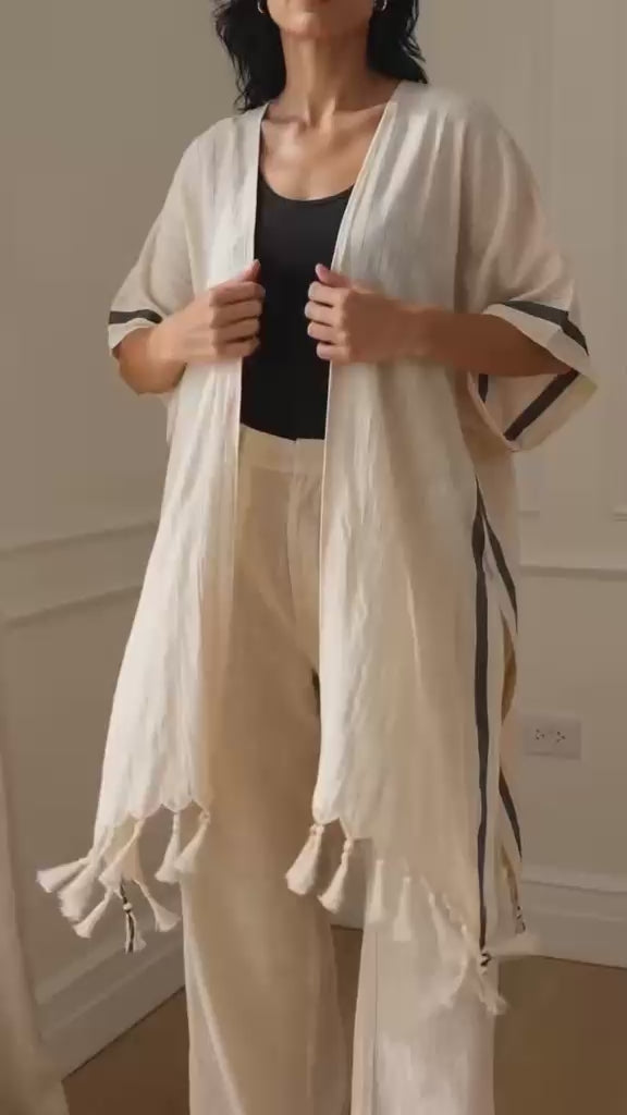 Asya Turkish cotton kimono shown in motion on a woman, featuring lightweight natural ivory woven cotton, an open-front relaxed silhouette, short sleeves with charcoal stripe details, high side splits, flowing drape, and hand-finished tassel fringe. Designed as a beach cover-up and versatile everyday layering piece.