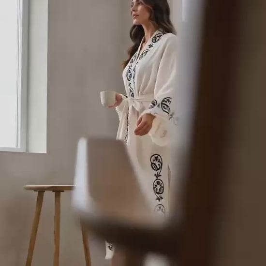 Woman wearing a handmade wood-block printed Turkish cotton kimono robe featuring colorful artisan motifs on soft natural cotton. The video shows the lightweight kimono in everyday wear, highlighting its hand-printed design, relaxed silhouette, wide sleeves, matching waist tie, breathable textured fabric, and comfortable drape. Artisan-crafted in Türkiye using traditional wood-block printing techniques, this natural cotton kimono robe is designed for lounging, slow mornings, travel, resort wear, beach days, 
