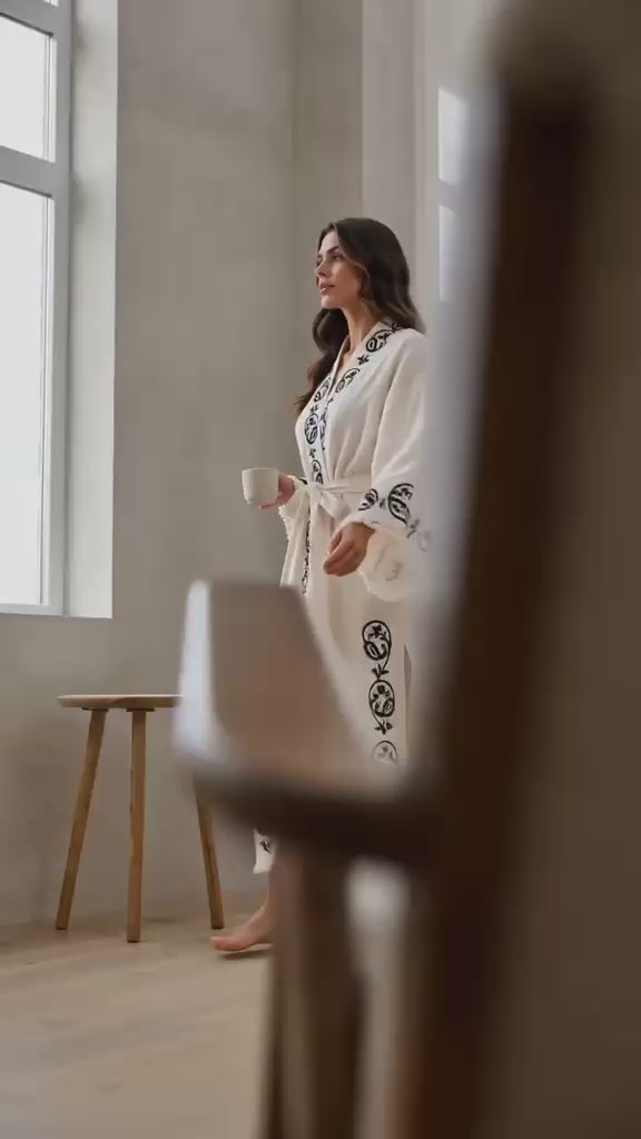Woman wearing a handmade wood-block printed Turkish cotton kimono robe featuring colorful artisan motifs on soft natural cotton. The video shows the lightweight kimono in everyday wear, highlighting its hand-printed design, relaxed silhouette, wide sleeves, matching waist tie, breathable textured fabric, and comfortable drape. Artisan-crafted in Türkiye using traditional wood-block printing techniques, this natural cotton kimono robe is designed for lounging, slow mornings, travel, resort wear, beach days, 