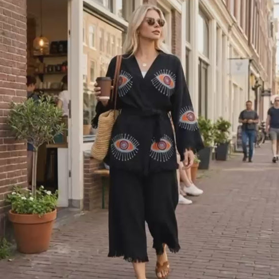 Black Turkish cotton evil eye kimono styled as an everyday streetwear look, featuring colorful eye motifs, relaxed wide sleeves, adjustable waist tie and fringe details. Woman walks through the city carrying coffee and a woven bag, showing the kimono’s relaxed fit and movement.