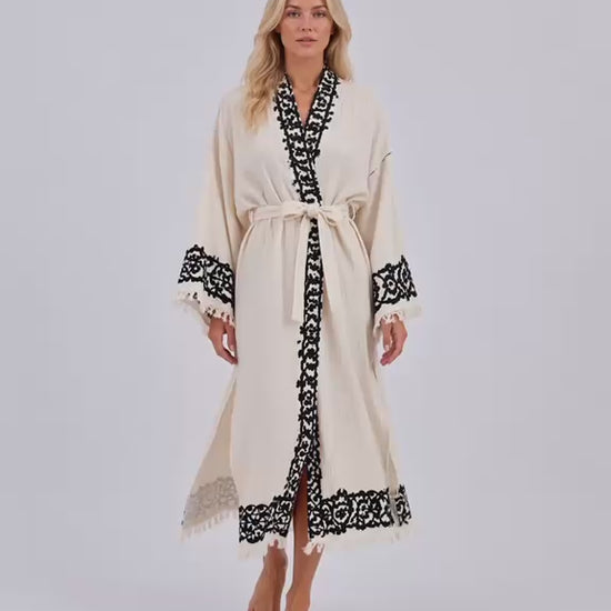 Woman wearing an ivory Turkish cotton kimono robe with contrasting black handmade wood-block printed motifs along the neckline, front opening, and wide sleeves. The video shows the lightweight, naturally textured cotton robe in soft movement inside a bright, sunlit interior, highlighting the relaxed silhouette, adjustable waist tie, fringe-trimmed cuffs, traditional artisan print details, and flowing drape. Handmade in Türkiye using traditional wood-block printing techniques, this breathable natural cotton 