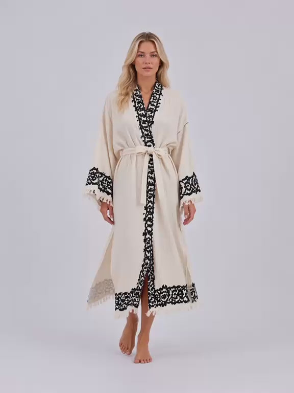 Woman wearing an ivory Turkish cotton kimono robe with contrasting black handmade wood-block printed motifs along the neckline, front opening, and wide sleeves. The video shows the lightweight, naturally textured cotton robe in soft movement inside a bright, sunlit interior, highlighting the relaxed silhouette, adjustable waist tie, fringe-trimmed cuffs, traditional artisan print details, and flowing drape. Handmade in Türkiye using traditional wood-block printing techniques, this breathable natural cotton 